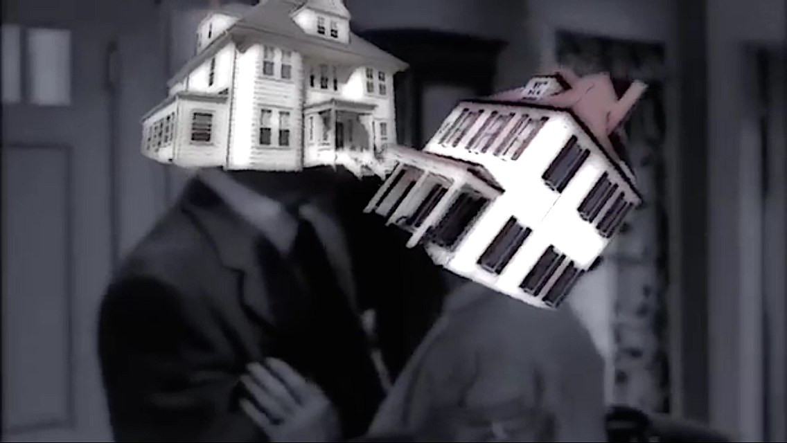 Black-and-white video still showing two figures wearing house-shaped structures.